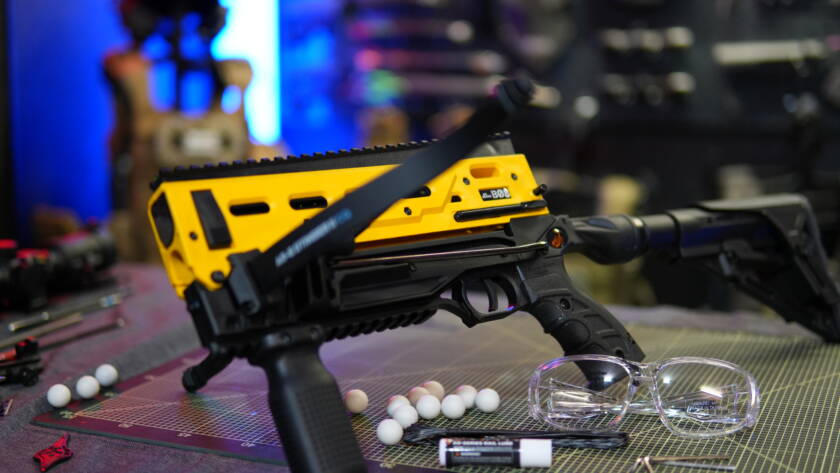 B08 less-lethal crossbow in yellow signal color designed for .68 caliber paintball ammunition, shown with safety glasses and projectiles on a work surface.