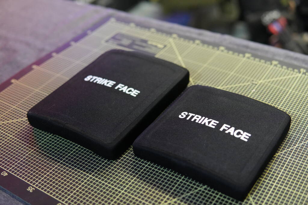 Two black ballistic side plates labeled “Strike Face” placed on a cutting mat – a smaller 6x6 plate on the right and a larger 6x8 side plate on the left.