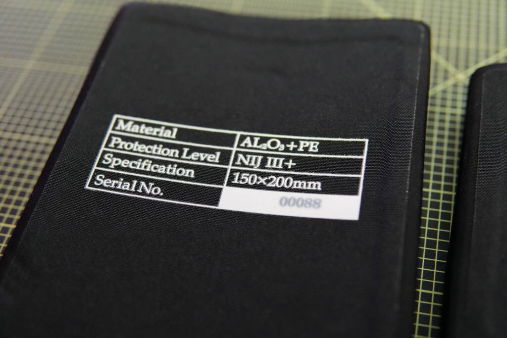Close-up of a black ballistic plate with a label showing material (Al₂O₃ + PE), protection level NIJ III+, and size 150 × 200 mm, placed on a cutting mat.