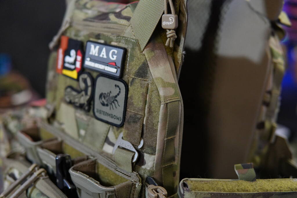 Close-up of an Agilite K-Zero SF plate carrier in camouflage with mounted magazine pouch, wide admin pouch, side plates, and several morale patches.