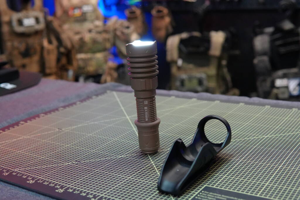 Olight Warrior X4 tactical flashlight in FDE with polymer holster, standing on a table with tactical gear in the background.