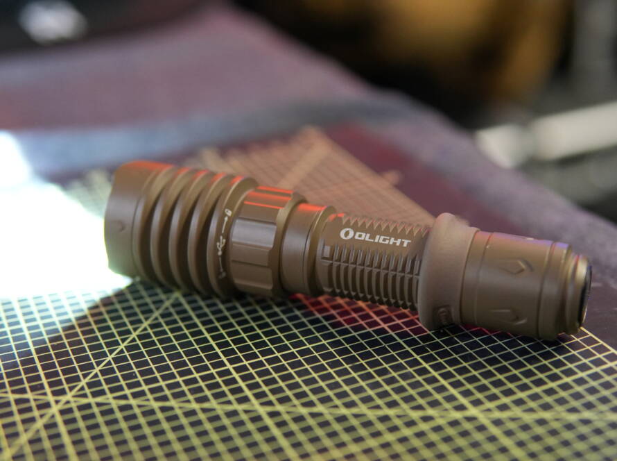 Olight Warrior X4 tactical flashlight turned on and lying on a workbench, emitting bright white light.
