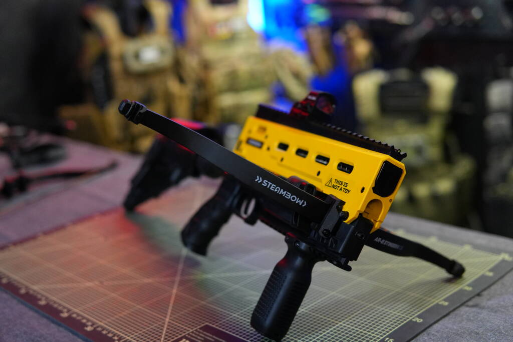 Steambow B08 less-lethal crossbow with bright yellow signal upper receiver on a workbench, contrasting strongly against tactical gear in the background.