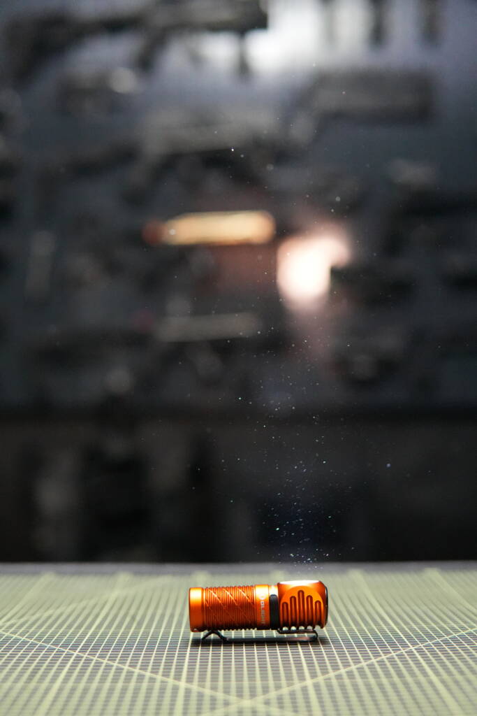 Olight Perun 3 Mini shining upward in a dark room, visible light beam illuminating dust particles in the air.
